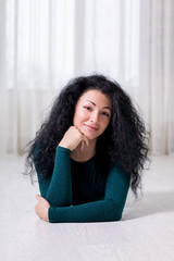 a portrait of a young happy girl with black curly brunette hair who lies on the white floor and holds her head with her hand, in a light interior and dark green pullover.
