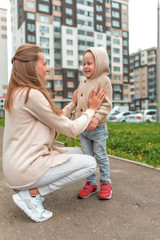 Young happy family, woman mom and little son boy. They relax in autumn on street, smile, walk, and rejoice. Casual warm clothes with a hood jumper. Background building road lawn.