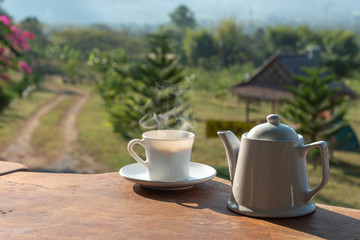 cup of coffee with white coffee mug on wooden table with scenery of mountain and field of plants in background