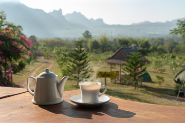 cup of coffee with white coffee mug on wooden table with scenery of mountain and field of plants in background