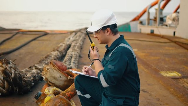 Marine chief officer or chief mate on deck of ship or vessel. He fills up ahts vessel checklist. Ship routine paperwork. He holds VHF walkie-talkie radio in hands.