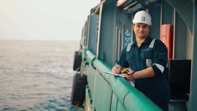 Marine chief officer or chief mate on deck of ship or vessel. He fills up ahts vessel checklist. Ship routine paperwork. He holds VHF walkie-talkie radio in hands.