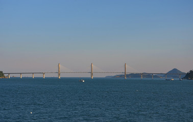The long span of a brdige asA bridge stretches over a dark blue bay. Diagonal struts radiate out from the middle pylons to the carriageway. Tree covered hills are in the distance.