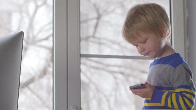 Cute blond boy is playing with smartphone, standing on his father work chair next to modern computer. child wants to quickly become adult and learn everything. Home education concept for lagging kids