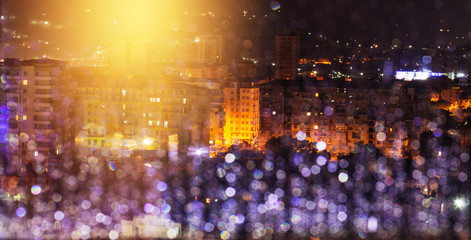 City in the rain at night. Window glass with raindrops defocused. Street lighting reflected and glare in the rain water in the foreground