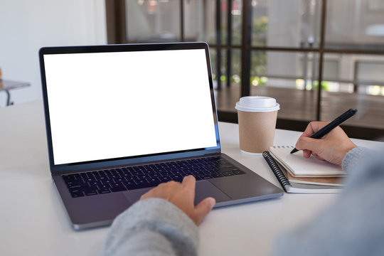 Mockup image of a woman using laptop with blank white desktop screen while writing on a notebook in office