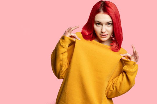 Beautiful young woman student touching warm hoodie with fingers looking at camera. Pretty stylish girl teenager with trendy red hair wearing yellow sweatshirt. Blank studio copy space background