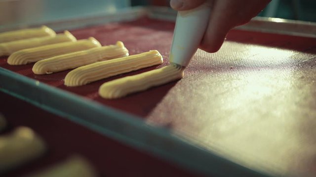 Preparing traditional French choux, profiteroles, squeezing from pipe on tray. Uncooked eclairs lay on pan in lines, looking tasty.