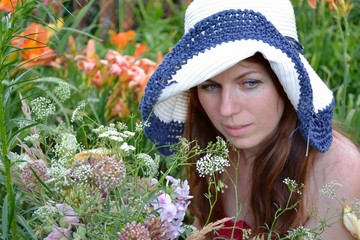 A young woman with long brown hair in a crimson sundress and a white hat with blue edges with a bouquet of wild flowers in her hand sitting in colorful lilies in the summer garden