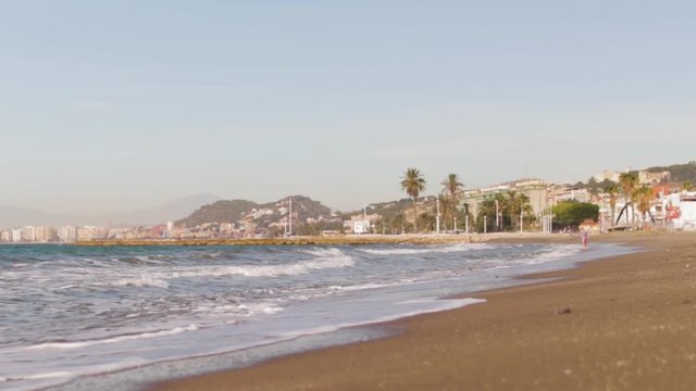 A very low angle Shot of crushing in waves in slow motion with palm trees, houses and mountain hills in the background