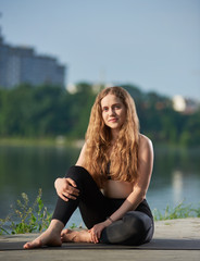 Fototapeta na wymiar Portrait of a beautiful young woman with long hair sitting on the mat near urban lake, smiling and looking at camera against blurred city view background