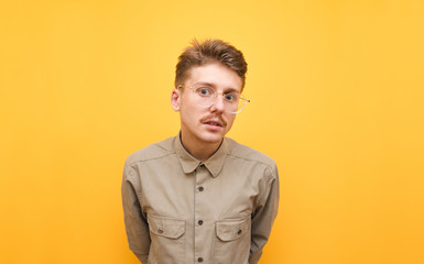 Obraz premium Portrait of a beautiful nerd with mustache and glasses stands against a yellow background and looks intently into the camera, wearing a beige shirt. Funny male student in shirt isolated on yellow
