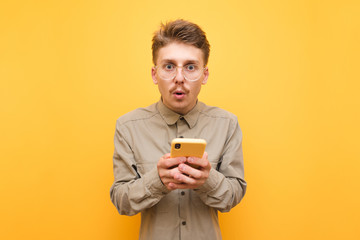 Surprised guy in glasses and mustache is standing on yellow background with smartphone in hands and looking into the camera.Portrait of surprised batman using smartphone isolated on yellow background
