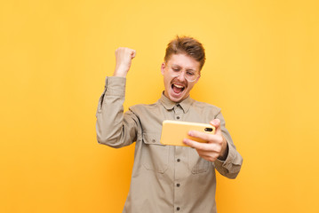 Portrait of happy guy in shirt and glasses looking at smartphone in hand and emotionally rejoicing....