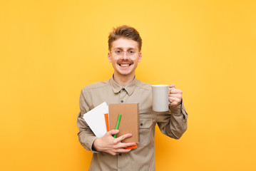 Happy student in shirt and glasses stands on yellow background with cup of hot drink in hand and exercise books, pen and books, looks into camera and smiles. Isolated. Portrait of happy nerd.