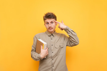Funny student with notebooks looks in camera and points finger at his head, wears glasses and a beige shirt.Nerd with books in his hand looks at the camera in amazement,points his finger his temple