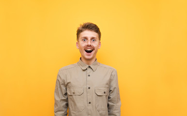 Portrait of funny joyful nerd in glasses and shirt on yellow background, looks into camera with funny face and smiles. A cheerful geek with a mustache looks at the camera in surprise. Isolated. - obrazy, fototapety, plakaty