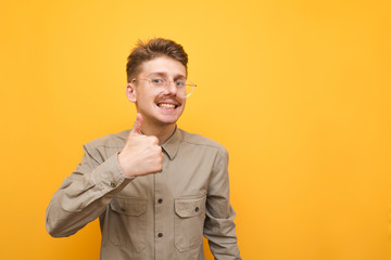 Happy nerd with mustache and glasses shows thumb up and looks into camera with happy face on yellow background. Geek in glasses and shirt shows a gesture of liking, lifted his thumb up.