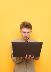 Funny nerd in glasses and mustache working on laptop on yellow background, looking shockingly at computer screen. Portrait of shocked guy with laptop in hands at work. Isolated. Copy space