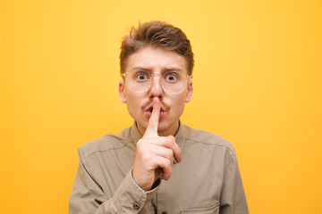 Closeup photo of surprised young man in glasses and mustache on yellow background, shows silence sign and with shocked face looks into camera.Emotional nerd put his finger to his lips and says shh