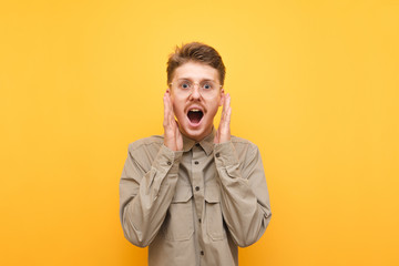 Close up portrait of shocked young man in shirt and glasses rejoicing, looking surprised at camera with mouth open. Joyful nerd with mustache shouts with happiness on yellow background. Isolated.