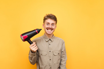 Portrait of funny young man in glasses and shirt on yellow background looking into camera with crazy face. Expressive fun guy uses a hairdryer. Isolation.
