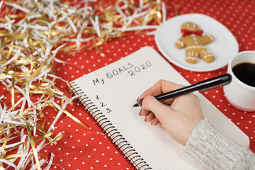 Female hands writing My Goals 2020 in a notebook. Gingerbread man, coffee, red background and...