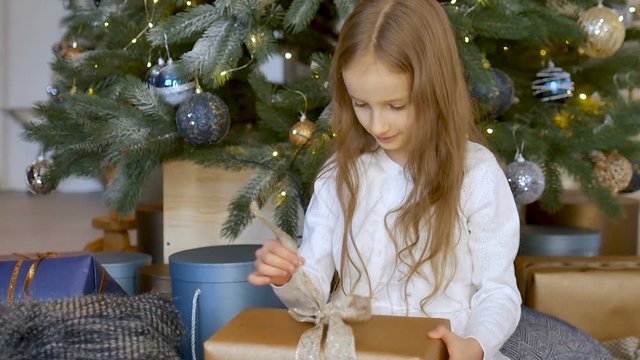 Cute blond girl has recieved a gift box wrapped in craft paper sitting near christmas tree, happy childhood and xmas celebration concepts