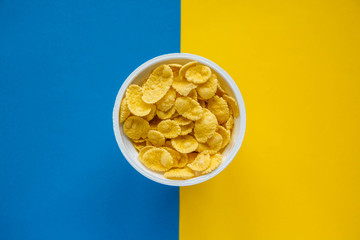 Cornflakes in white bowl on blue and yellow background. Top view. Copy, empty space for text