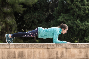 Side view of young female athlete with wireless earphones doing abdominal plank on top of a park wall.