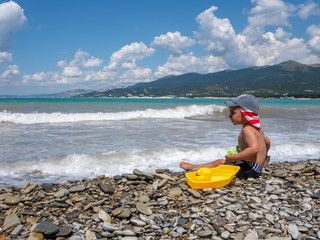 A little boy with panama hat plays on the beach by the sea with a yellow toy ship and green shoulder blade