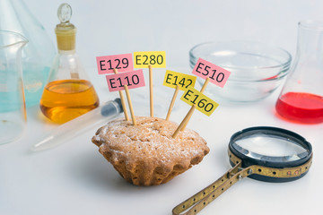 A cupcake decorated with name plates of additives E, test tubes and a magnifier stand nearby. Food laboratory. Close up on a white table.