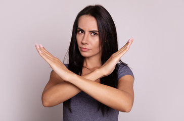 Please, do not! Close-up photo of a young serious dark-haired girl in a grey t-shirt, who is looking in the camera and showing a cross with her arms.
