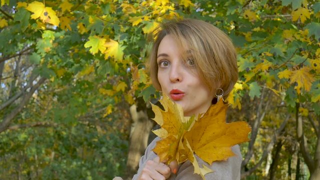 Emotional attractive young woman with blonde short hair and biege dress holding a bouquet of autumn leaves above her head and posing in a beautiful park outdoors