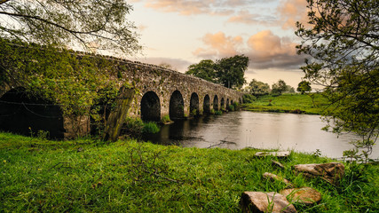 Old 12th century stone arch bridge over a river, rocks on first plane. Green fields and trees. Dramatic sky sunset. Count Meath, Ireland