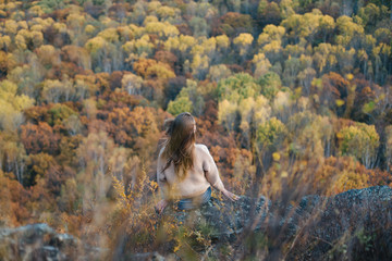 Young woman sitting on cliff's edge, looking at beautiful autumn forest.