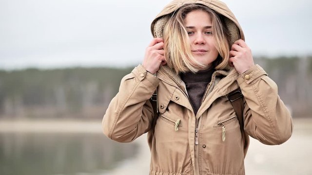 portrait of a young smiling girl with short blonde hair standing against the sea, looking into the distance, putting on a hood of a warm jacket and going into the distance along the shore