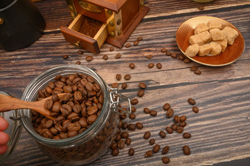 The girl's hand takes a wooden spoon of coffee beans from a glass jar, a coffee grinder, pieces of brown sugar on a wooden background. Close up.