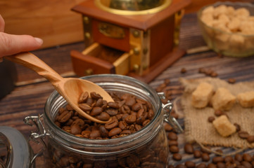 The girl's hand takes a wooden spoon of coffee beans from a glass jar, a coffee grinder, pieces of brown sugar on a wooden background. Close up.