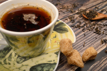 A Cup of black tea, tea leaves in a wooden spoon, pieces of brown sugar on a wooden background. Close up.