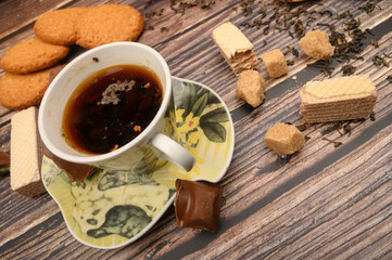 A Cup of black tea, tea leaves, pieces of brown sugar, oatmeal cookies, waffles, pieces of chocolate on a wooden background. Close up.