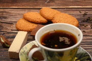A Cup of black tea, tea leaves, pieces of brown sugar, oatmeal cookies, waffles, pieces of chocolate on a wooden background. Close up.