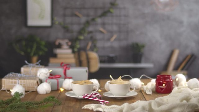 Dolly shot of two tasty cups of coffee cappuccino with sweet cream. Wooden table with Christmas presents, string of lights, candy canes, a warm white woolen blanket and a branch of spruce. Home.