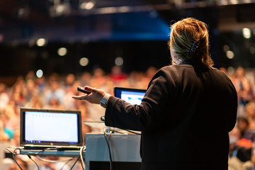 Female speaker giving a talk on corporate business conference. Unrecognizable people in audience at...