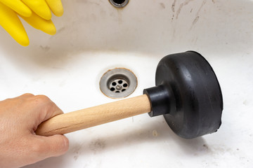 Blocked sewer, clogged wash bowl, basin drain, yellow rubber gloves and a hand holding a plunger in...