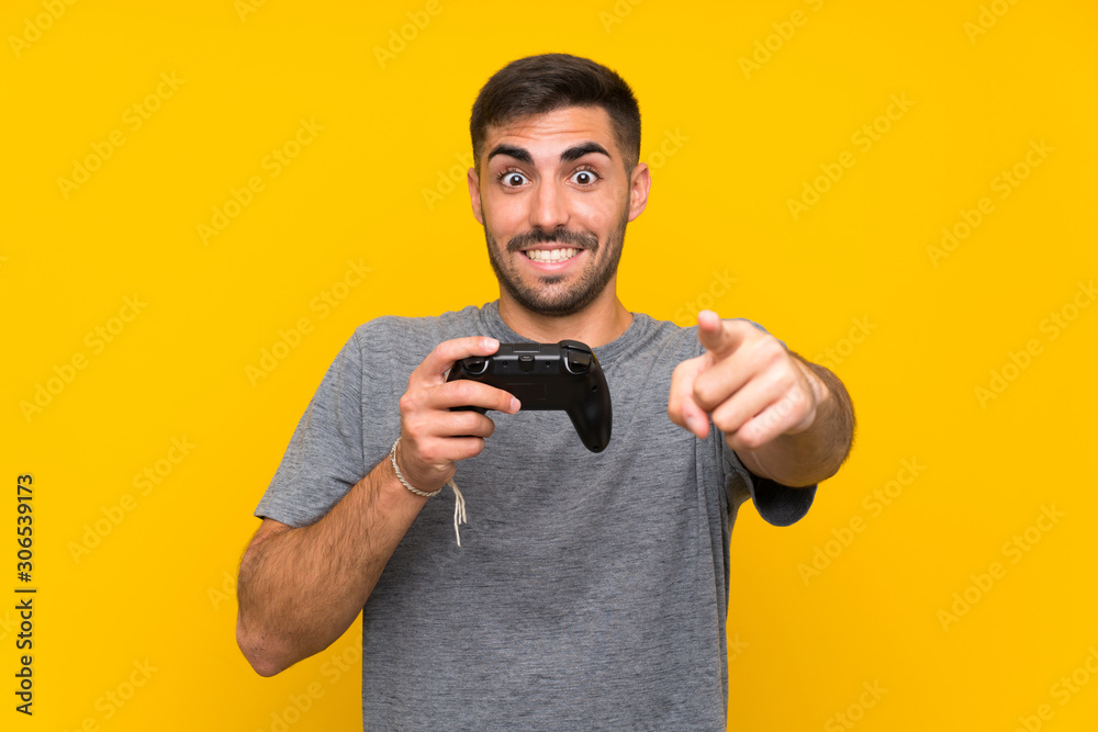 Wall mural Young handsome man playing with a video game controller over isolated yellow background