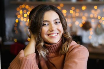 Close-up portrait of a beautiful attractive cute girl with short curly brunette hair with a beautiful smile in a warm cozy sweater smiling and laughing on a dark background of garlands and bokeh.