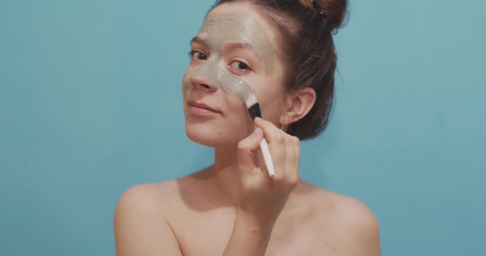 Portrait of attractive woman applying cleansing clay mask on face using make up brush indoors. Close up of Caucasian girl using facial cosmetics on blue background. Skin care treatment wellness