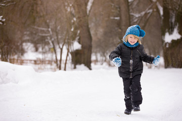 Kid boy runs on a snowy road in a park. Cheerful child with blond hair in blue mittens, a hat and a black jacket. Activities with children outdoors on a frosty winter day.
