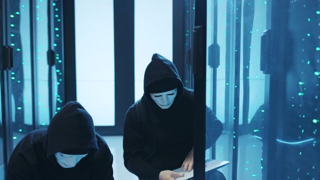 Professional team of hackers steaking into database server room. Malicious masked men connecting laptop to server cabinet to steal big data information. Cybercrime.